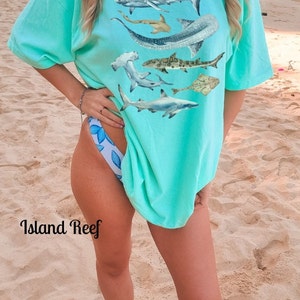 May include: A turquoise blue t-shirt with a graphic print of various sharks and rays. The shirt is oversized and has a relaxed fit. The model is wearing the shirt with a white and blue patterned bikini bottom. The text "Island Reef" is visible in the bottom left corner of the image.