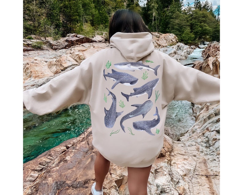 May include: A beige hoodie with a graphic print of multiple whale sharks on the back. The hoodie has a drawstring hood and a kangaroo pocket.