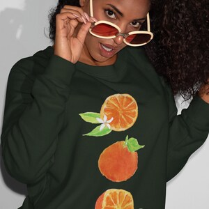May include: A dark green sweatshirt with a graphic of three orange slices and white blossoms. The oranges are stacked vertically and have a watercolor-like appearance.