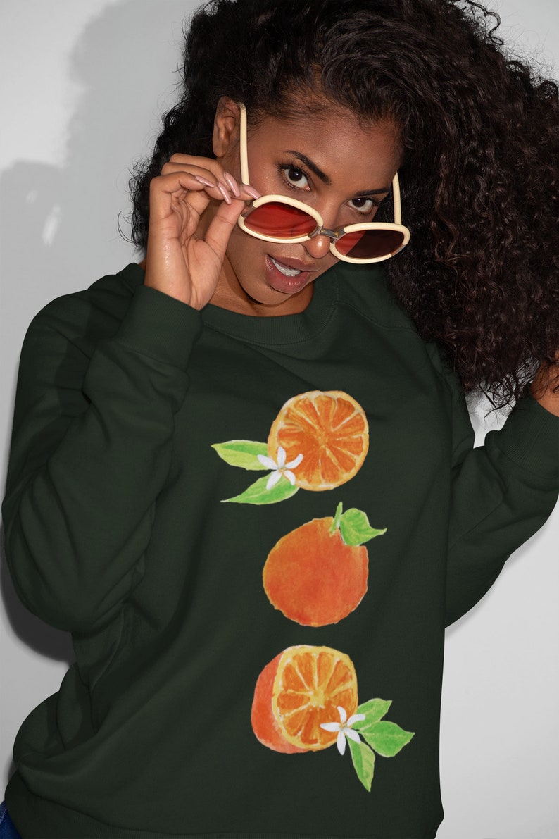 May include: A dark green sweatshirt with a graphic of three orange slices and white blossoms. The oranges are stacked vertically and have a watercolor-like appearance.