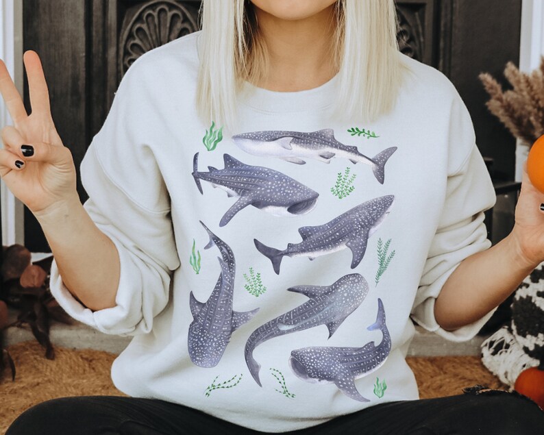 May include: A white sweatshirt with a graphic print of multiple whale sharks and seaweed. The whale sharks are gray with white spots and the seaweed is green.