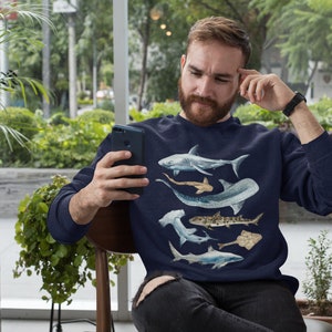 May include: A navy blue sweatshirt with a graphic print of various sharks. The sweatshirt features a white shark, a hammerhead shark, a nurse shark, a sand tiger shark, and a stingray.