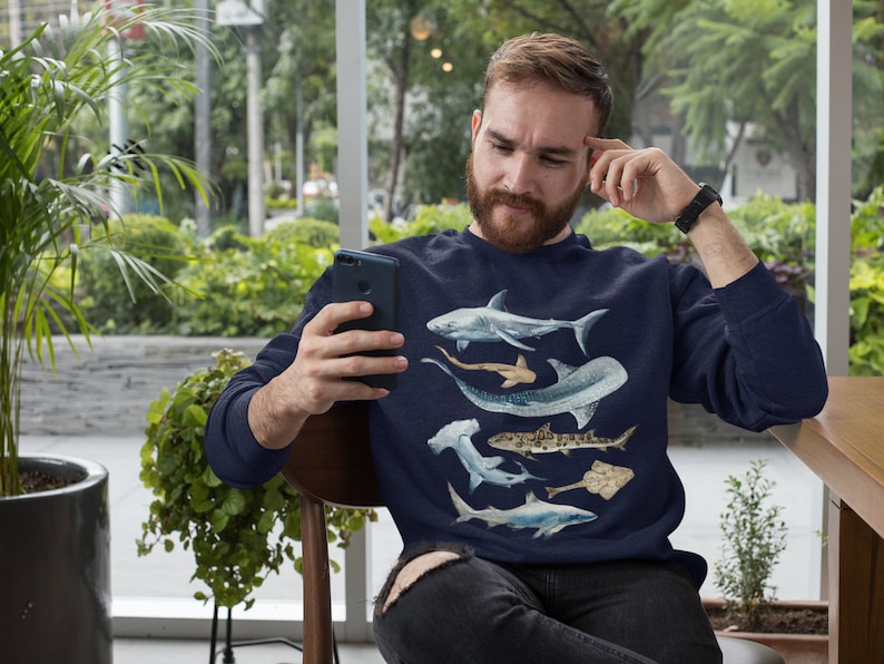 May include: A navy blue sweatshirt with a graphic print of various sharks. The sweatshirt features a white shark, a hammerhead shark, a nurse shark, a sand tiger shark, and a stingray.