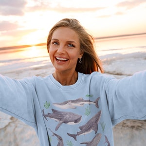 May include: A woman wearing a light blue sweatshirt with a print of whale sharks and seaweed. She is smiling and taking a selfie on a beach at sunset.