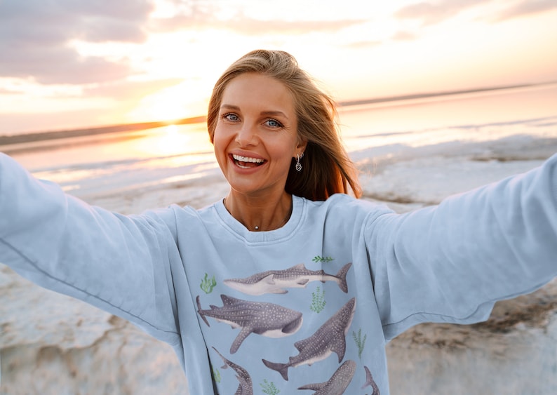 May include: A woman wearing a light blue sweatshirt with a print of whale sharks and seaweed. She is smiling and taking a selfie on a beach at sunset.