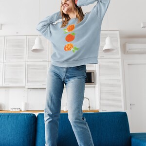 May include: A woman wearing a light blue sweatshirt with a graphic of three orange slices and white blossoms. She is standing on a blue couch with her arms raised above her head.