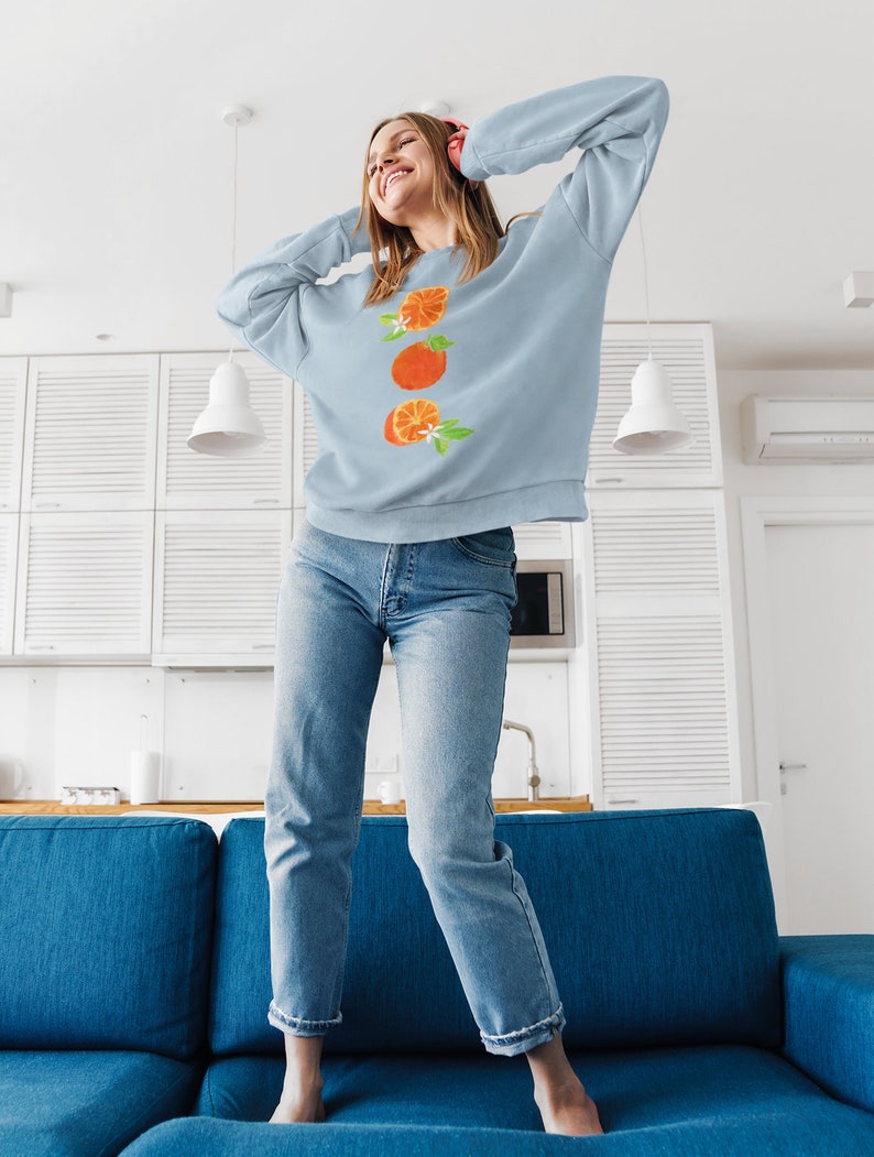 May include: A woman wearing a light blue sweatshirt with a graphic of three orange slices and white blossoms. She is standing on a blue couch with her arms raised above her head.