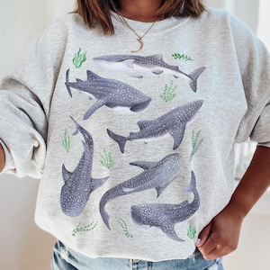May include: A gray sweatshirt with a graphic print of multiple whale sharks in various poses. The sharks are black and white with white spots. The background is a light gray with green seaweed.