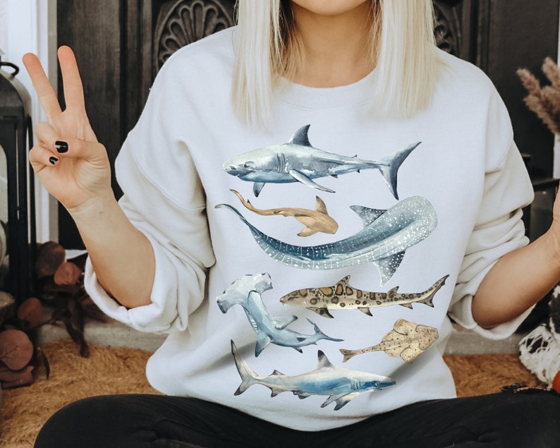 May include: White sweatshirt featuring a watercolour design of various sharks and rays. The design includes a large great white shark, a hammerhead shark, a whale shark, a stingray, and a smaller shark.