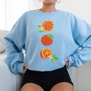 May include: A light blue sweatshirt with a graphic of three orange slices and white blossoms. The sweatshirt has a crew neck and long sleeves.