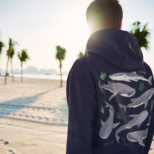 May include: A navy blue hoodie featuring a graphic print of several whale sharks and seaweed on the back. The person wearing the hoodie is standing on a beach with palm trees in the background.