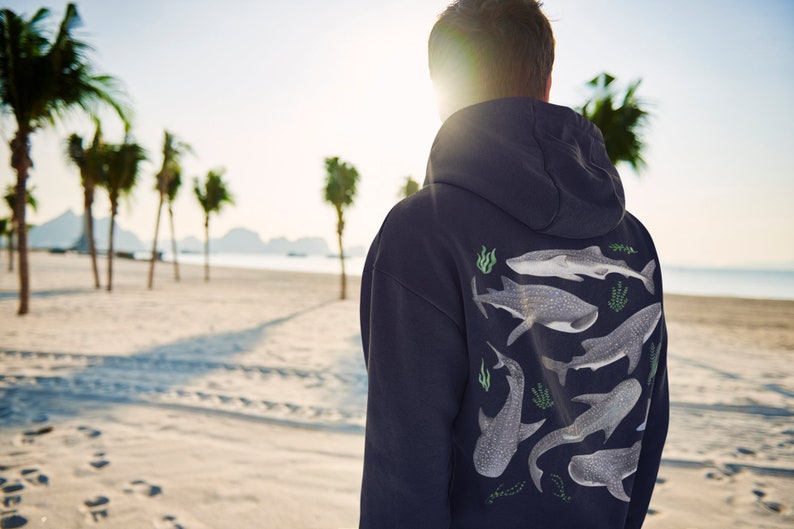May include: A navy blue hoodie featuring a graphic print of several whale sharks and seaweed on the back. The person wearing the hoodie is standing on a beach with palm trees in the background.