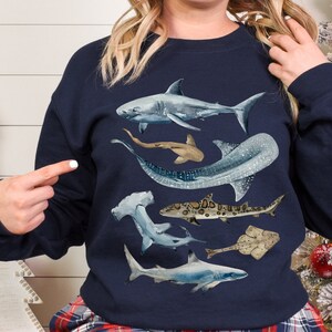 May include: A navy blue sweatshirt featuring a watercolour design of various sharks and rays. The design includes a hammerhead shark, a whale shark, a grey reef shark, a stingray, and a sawfish.
