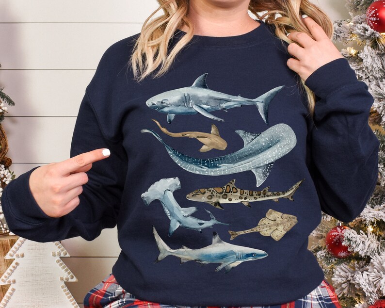 May include: A navy blue sweatshirt featuring a watercolour design of various sharks and rays. The design includes a hammerhead shark, a whale shark, a grey reef shark, a stingray, and a sawfish.