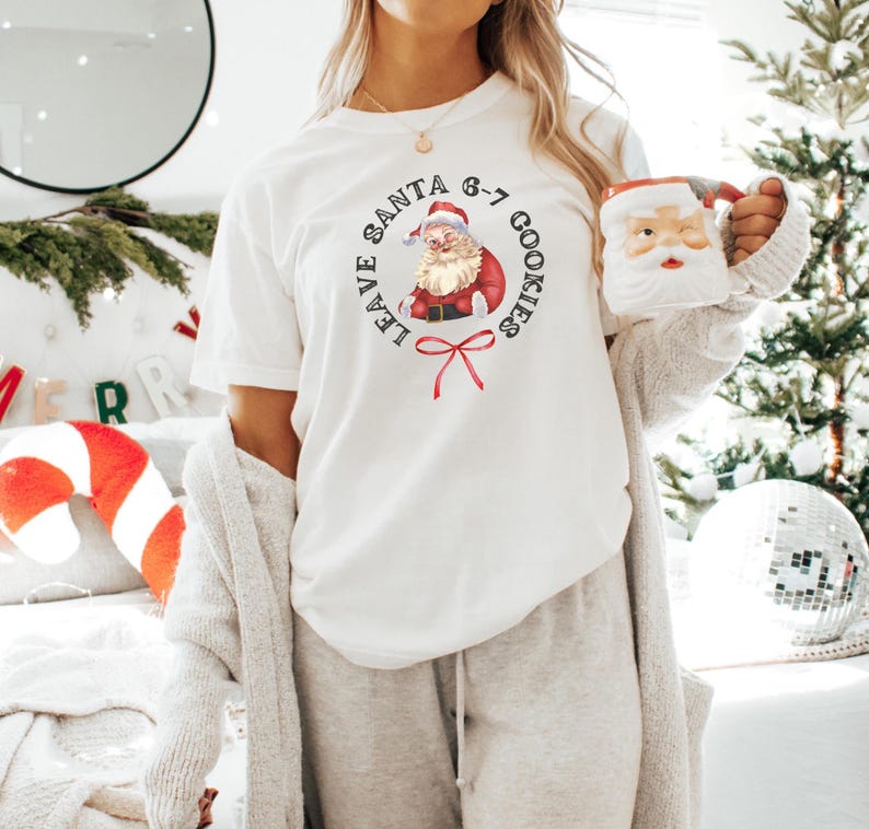 May include: White t-shirt with a circular graphic featuring Santa Claus and the text "LEAVE SANTA 6-7 COOKIES". The Santa graphic is in red, white, and black. A Santa-themed mug is held in the hand.