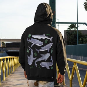 May include: A black hoodie featuring a graphic print of several whale sharks in shades of grey and blue, with green seaweed accents. The hoodie has a drawstring hood and a kangaroo pocket.