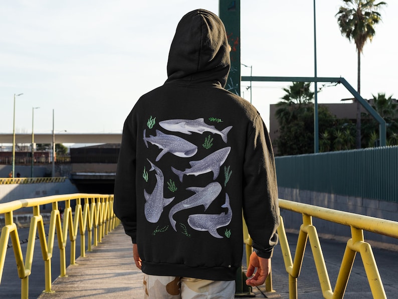 May include: A black hoodie featuring a graphic print of several whale sharks in shades of grey and blue, with green seaweed accents. The hoodie has a drawstring hood and a kangaroo pocket.