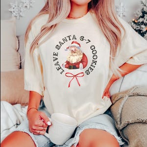 May include: Cream-colored t-shirt featuring a vintage Santa Claus graphic with the text "LEAVE SANTA 6-7 COOKIES". The Santa illustration is in a circular design with a red bow at the bottom. The shirt is worn with denim shorts.