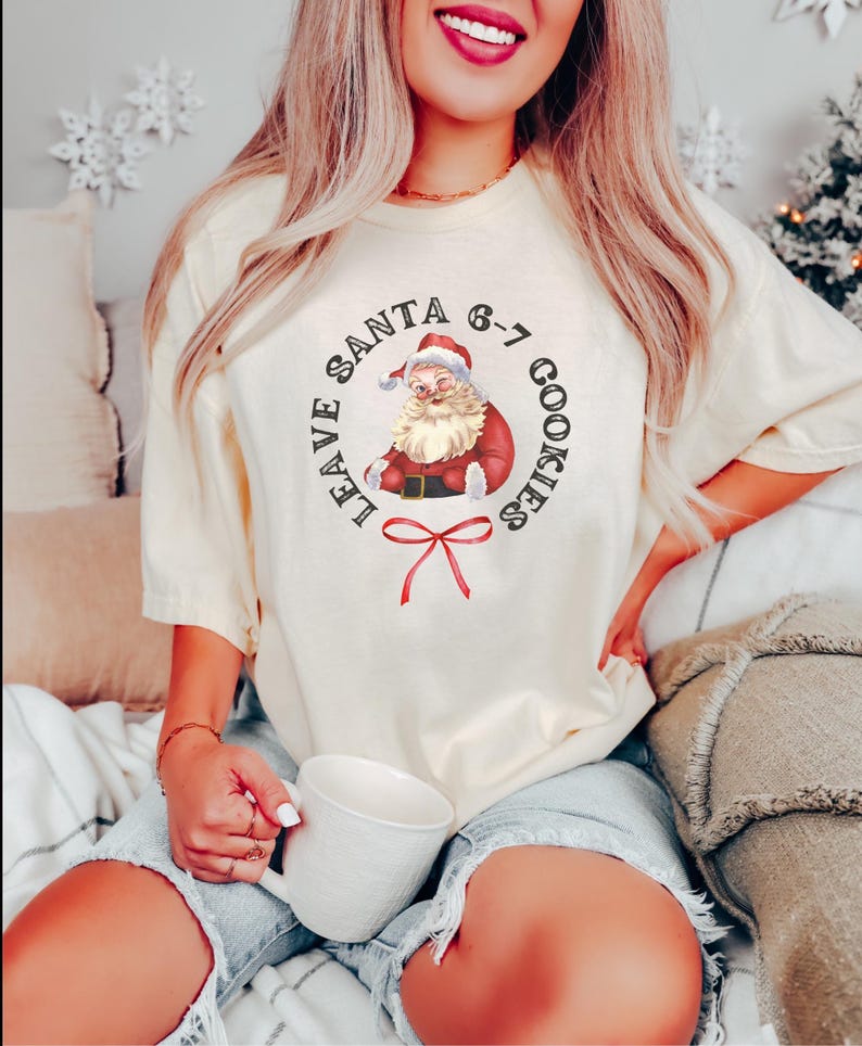 May include: Cream-colored t-shirt featuring a vintage Santa Claus graphic with the text "LEAVE SANTA 6-7 COOKIES". The Santa illustration is in a circular design with a red bow at the bottom. The shirt is worn with denim shorts.