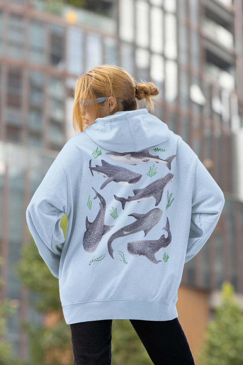May include: A light blue hoodie featuring a graphic print of several whale sharks in different positions. The sharks are surrounded by green seaweed.