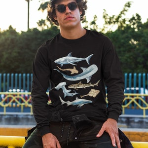 May include: A black long-sleeved shirt featuring a graphic print of five different types of sharks. The sharks are in shades of blue and grey, and are arranged in a layered pattern. The shirt is being worn by a person with a black cap and sunglasses.