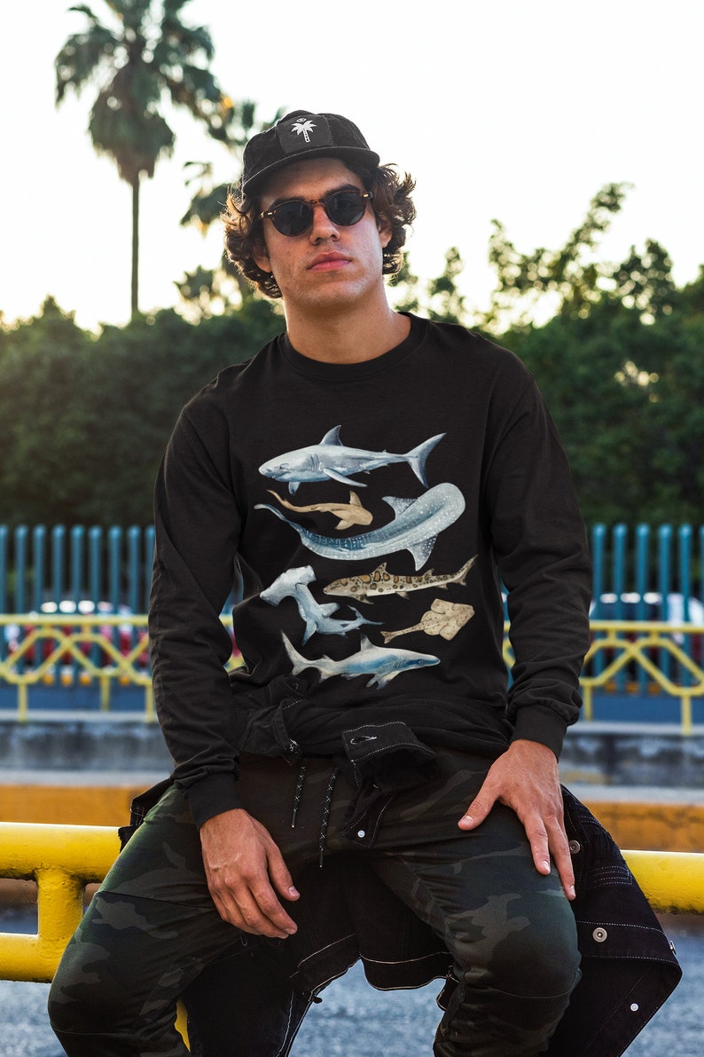 May include: A black long-sleeved shirt featuring a graphic print of five different types of sharks. The sharks are in shades of blue and grey, and are arranged in a layered pattern. The shirt is being worn by a person with a black cap and sunglasses.