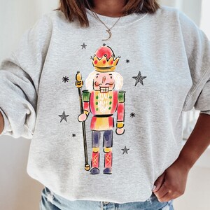 May include: A gray sweatshirt with a colorful illustration of a nutcracker holding a staff. The nutcracker is wearing a red and gold suit with a crown. The background is a light gray with stars.