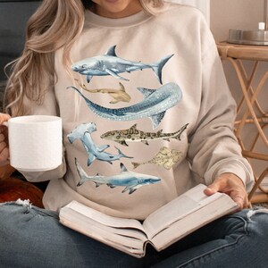 May include: A beige sweatshirt featuring a watercolour design of various sharks, including a hammerhead, a whale shark, and a great white shark.