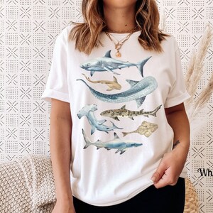 May include: White t-shirt with a watercolor design of various sharks, including a hammerhead, a whale shark, and a great white shark.