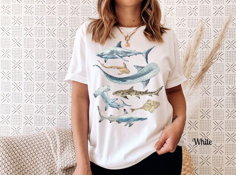 May include: White t-shirt with a watercolor design of various sharks, including a hammerhead, a whale shark, and a great white shark.