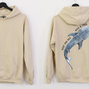 May include: A beige hoodie with a pocket on the front. The back of the hoodie features a blue and white whale shark with the text "Keep the Sea Wild & Free".