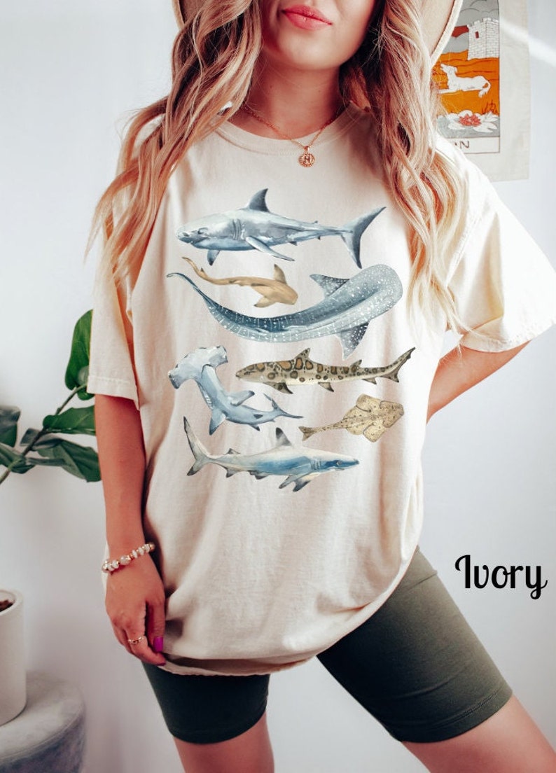 May include: A white t-shirt with a graphic print of various sharks, including a hammerhead, a whale shark, and a great white shark. The shirt is shown on a model wearing green shorts. The text "Ivory" is visible in the lower right corner of the image.