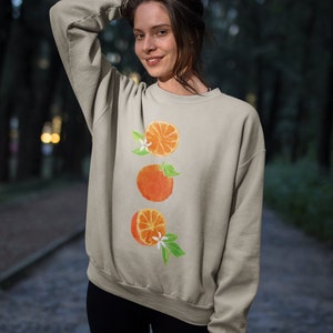May include: A light beige sweatshirt with a watercolor design of three orange slices with green leaves and white flowers.