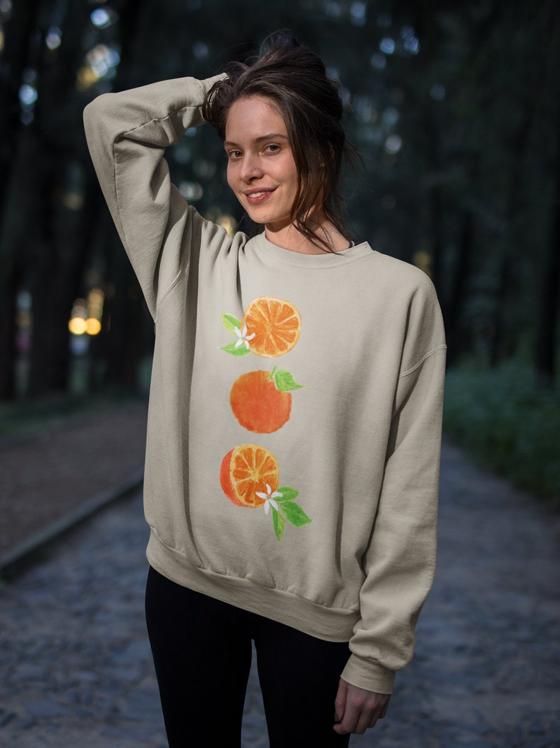 May include: A light beige sweatshirt with a watercolor design of three orange slices with green leaves and white flowers.