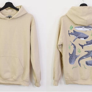 May include: A beige hoodie with a front pocket. The back of the hoodie features a design of multiple whale sharks in a light blue and grey colour scheme with green seaweed.
