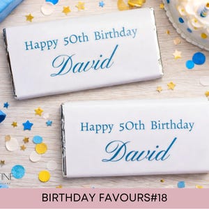 May include: Two rectangular chocolate bars wrapped in white paper with blue text that reads Happy 50th Birthday David. The wrappers have silver foil edges and are displayed among blue and gold confetti, a party hat, and a birthday cake.