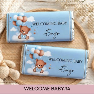 May include: Two rectangular chocolate bars wrapped in light blue paper featuring a brown teddy bear holding a cluster of blue and tan balloons. The text WELCOMING BABY and the name Enzo are printed in black on the front of each wrapper.