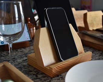 May include: A wooden phone stand with a natural finish. The stand has a slot for a phone to be placed in a vertical position. A black smartphone is in the stand.
