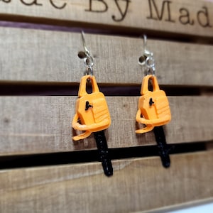 Puede incluir: Un par de pendientes originales con colgantes de motosierra naranja y negra en miniatura. Los pendientes cuelgan de ganchos de color plateado. El fondo es una caja de madera con las palabras "Made By Mada" grabadas.