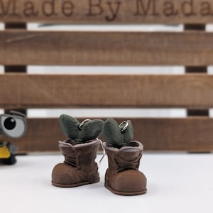 Puede incluir: Pendientes de bota marrón con detalles de plantas verdes. Las botas tienen cordones y un ribete de piel marrón.