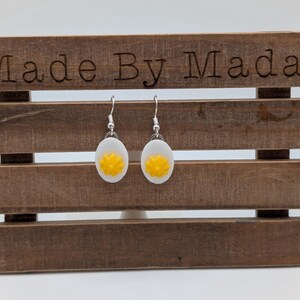 May include: A pair of silver earrings with white and yellow egg-shaped charms. The earrings are displayed on a wooden stand with the text "Made By Mada" on the top.