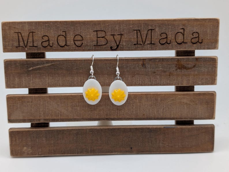 May include: A pair of silver earrings with white and yellow egg-shaped charms. The earrings are displayed on a wooden stand with the text "Made By Mada" on the top.