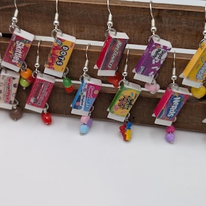 May include: A set of candy-themed earrings featuring various popular candy brands, including Skittles, Whoppers, Sweetarts, and Nerds. Each earring features a miniature box of candy with a colorful bead dangling below.