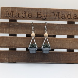May include: A pair of dangle earrings featuring a small potted succulent plant hanging from a white string with a wooden bead. The earrings are displayed on a wooden shelf with the text "Made By Mada" engraved on the top.