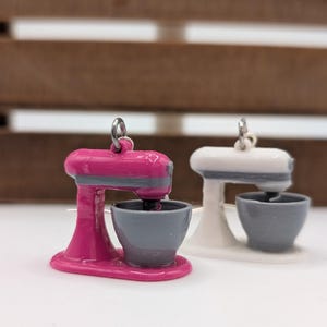 May include: A pair of pink and white stand mixer earrings. The earrings are made of plastic and have a silver hook.