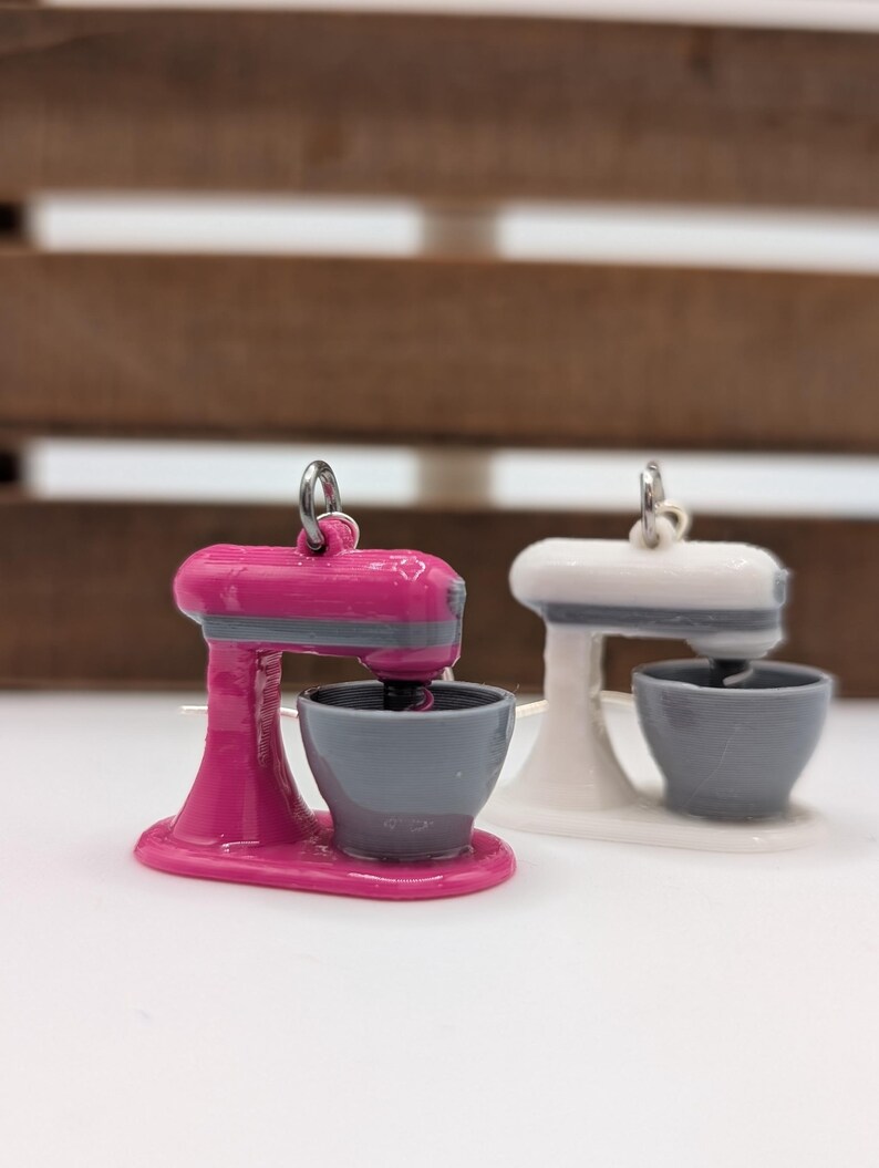 May include: A pair of pink and white stand mixer earrings. The earrings are made of plastic and have a silver hook.