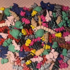 May include: A vibrant collection of miniature sculpted creatures in diverse forms and hues, such as pink axolotls, green turtles, and blue sharks. Other colours include yellow, red, and grey. The assortment is scattered on a pink floral background.
