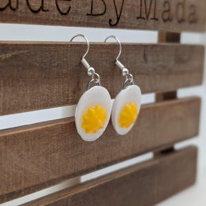 May include: A pair of dangle earrings featuring white and yellow egg-shaped charms. The earrings are made of polymer clay and have silver hooks.