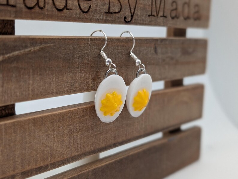 May include: A pair of dangle earrings featuring white and yellow egg-shaped charms. The earrings are made of polymer clay and have silver hooks.