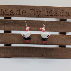 May include: Handmade earrings with a white cup design, red and white striped straw, black base, and red accents. The earrings are displayed on a wooden stand with the text "Made By Mada".
